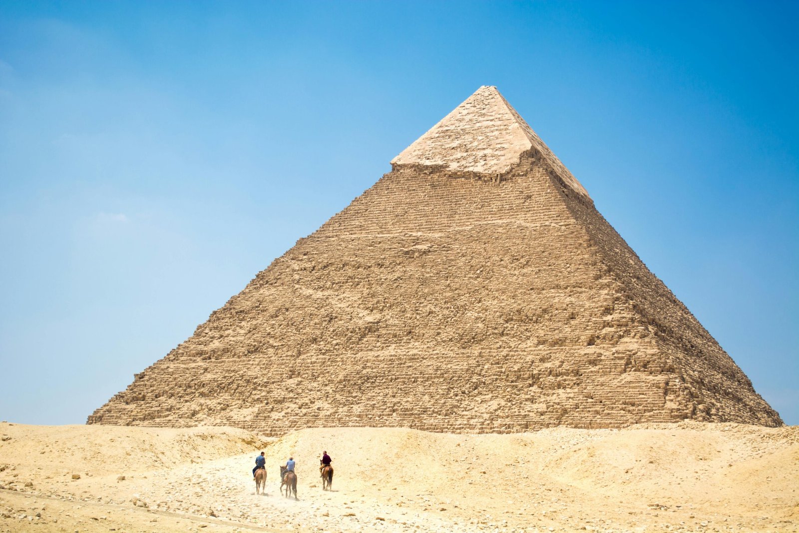 A panoramic view of the Great Pyramid of Giza under a bright blue sky, with a few tourists exploring the base of the ancient structure