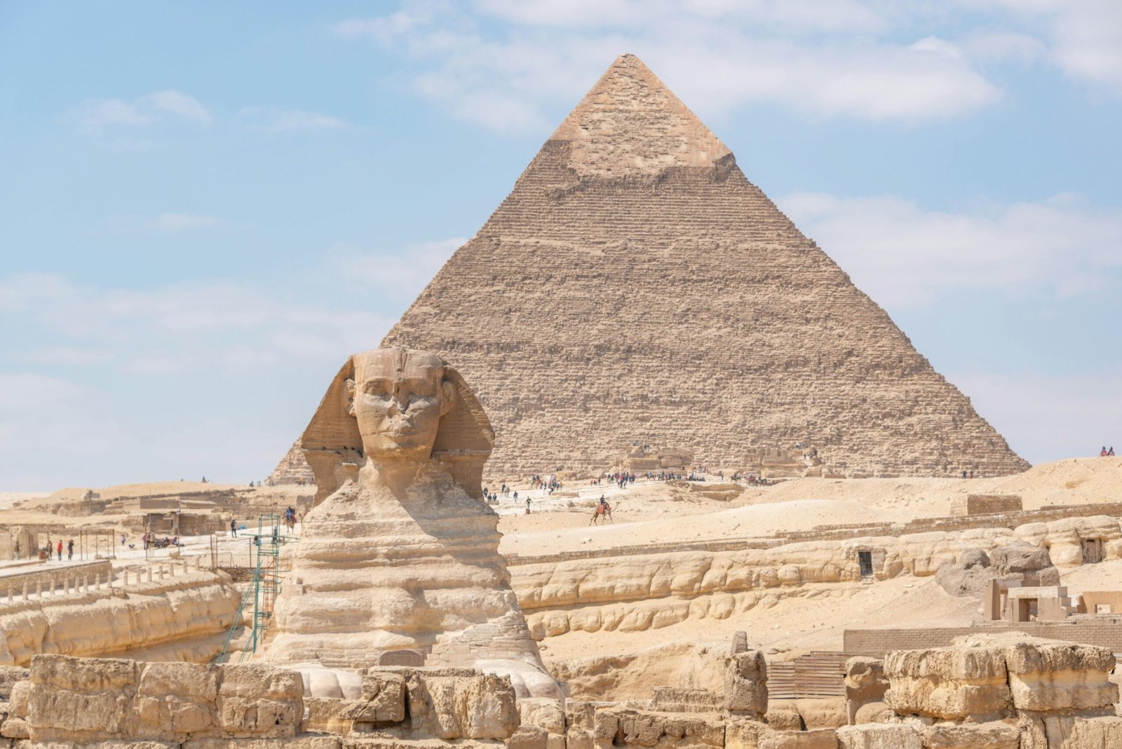 A panoramic view of The Great Pyramid of Khafre, standing tall with its smooth apex visible, under a bright blue sky, with tourists exploring its base.